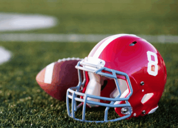 A red football helmet with the number 8 on it and a football lying on a green field with white yard line markings, representing the Aer Lingus College Football Classic.
