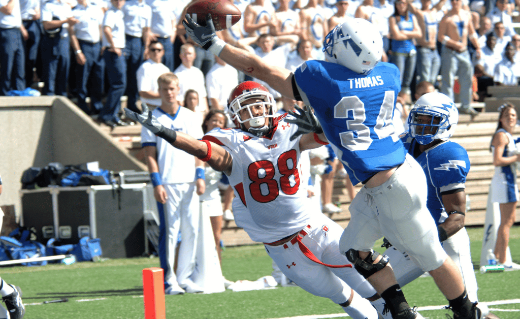 A football player in a blue jersey with the number 34 and the name Thomas is catching a football mid-air while being closely defended by a player in a white jersey with the number 88. The scene takes place on a football field with spectators in the background, representing the Aer Lingus College Football Classic.