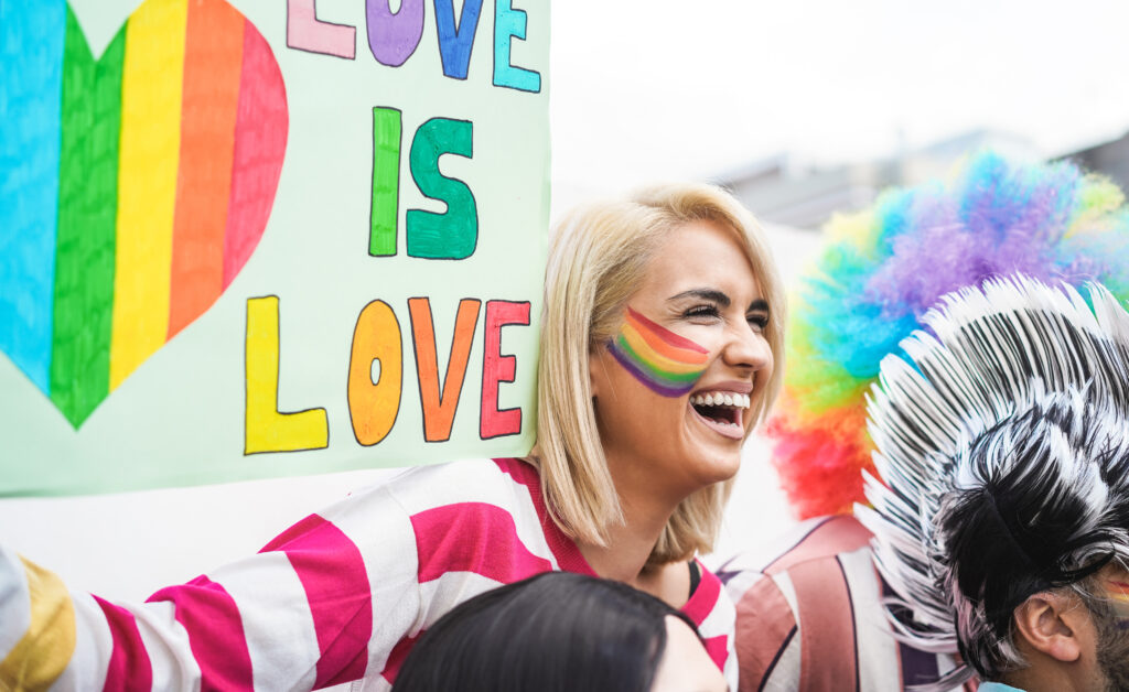 A person with a blurred face stands in front of a colorful sign that reads "LOVE IS LOVE" with the word "LOVE" written in rainbow colors. Another person next to them is wearing a headdress with feathers. Dublin Pride