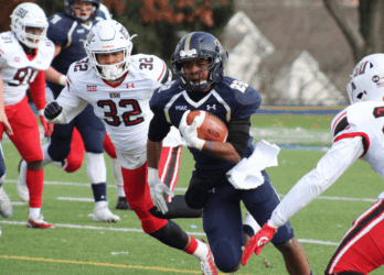 NFL Weekend in Dublin: A football player in a navy blue uniform is running with the ball, pursued by players in white and red uniforms on a green field. The background shows blurred trees and buildings.