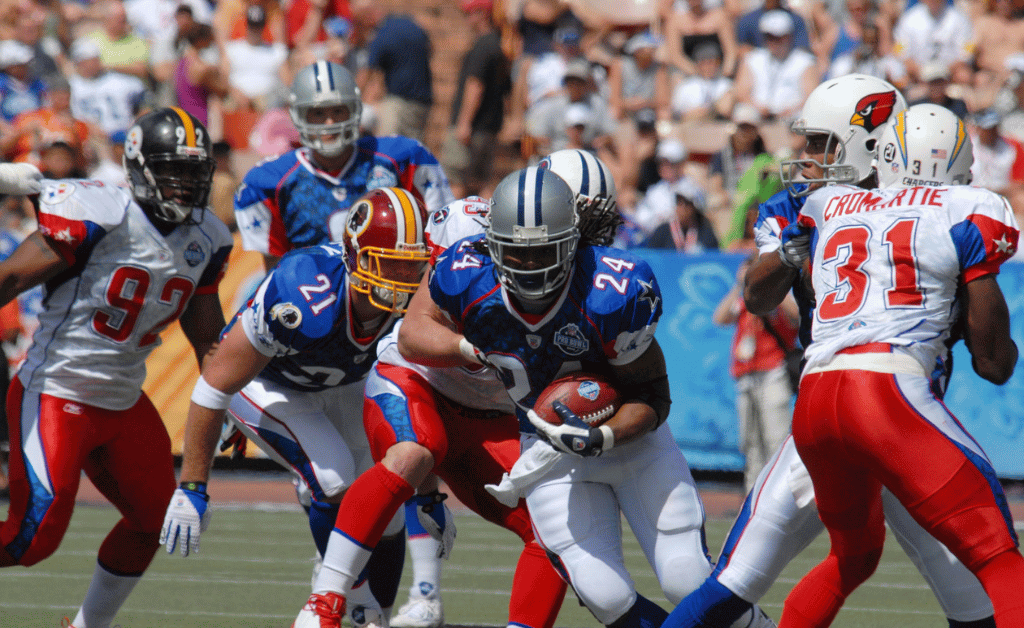 NFL weekend in Dublin: A football player in a blue jersey with the number 24 is running with the ball, while other players attempt to tackle or block him. The background shows a crowd of spectators.