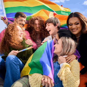 Dublin Pride, a group of people sitting together, holding and waving rainbow flags. The background shows a clear blue sky with some clouds.