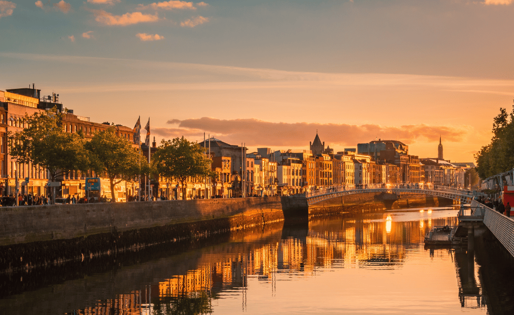 A scenic view of the River Liffey in Dublin, Ireland, during sunset. The river is flanked by historic buildings and trees on both sides. A bridge spans the river in the middle of the image, and the water reflects the warm hues of the setting sun and surrounding architecture. This picturesque scene is near a hotel near Grafton Street.