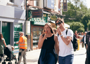 A busy scene on Grafton Street, with several people walking. Two individuals in the foreground have their faces blurred and are walking side by side, one holding a phone. There are various shops and buildings in the background, including a store with green signage that reads &#039;Celtic&#039; and another shop with a sign that says &#039;TO LET.&#039; Other pedestrians can be seen in the background, including someone wearing an orange safety vest. The weather appears to be sunny.