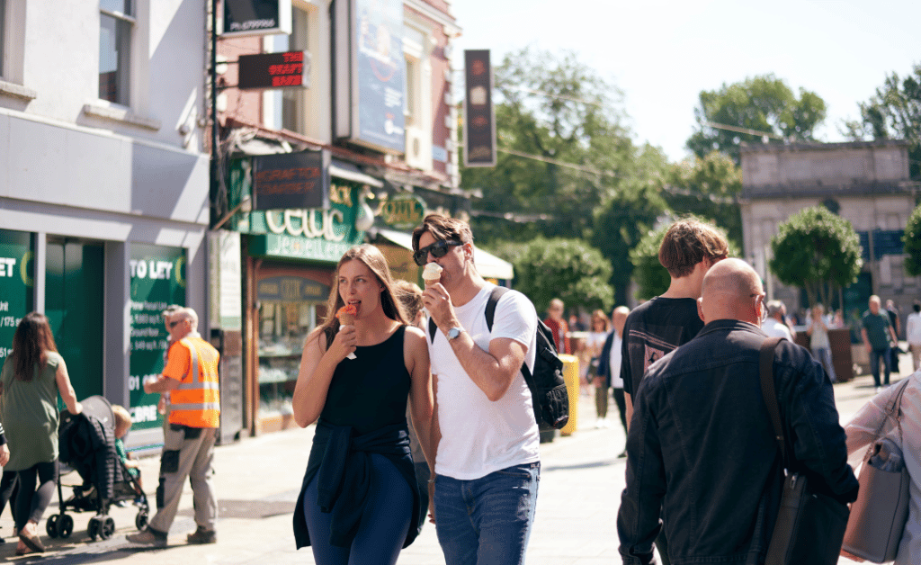 A busy scene on Grafton Street, with several people walking. Two individuals in the foreground have their faces blurred and are walking side by side, one holding a phone. There are various shops and buildings in the background, including a store with green signage that reads 'Celtic' and another shop with a sign that says 'TO LET.' Other pedestrians can be seen in the background, including someone wearing an orange safety vest. The weather appears to be sunny.