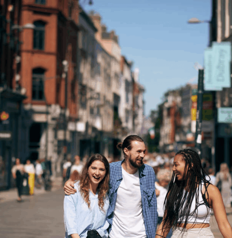 A sunny day on Grafton Street, a busy shopping street in Dublin, Ireland. The image shows three people walking down the street with their faces blurred. They are surrounded by various shops and buildings, including recognizable brands like Levi's, Brown Thomas, Zara, Disney. The street is bustling with pedestrians in the background.