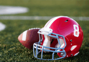 A red football helmet with the number 8 on it and a football lying on a green field with white yard line markings, representing the Aer Lingus College Football Classic.