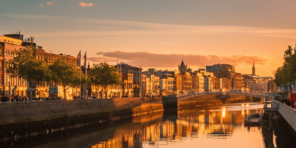 A scenic view of the River Liffey in Dublin, Ireland, during sunset. The river is flanked by historic buildings and trees on both sides. A bridge spans the river in the middle of the image, and the water reflects the warm hues of the setting sun and surrounding architecture. This picturesque scene is near a hotel near Grafton Street.