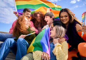 Dublin Pride, a group of people sitting together, holding and waving rainbow flags. The background shows a clear blue sky with some clouds.