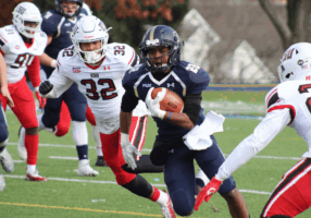 NFL Weekend in Dublin: A football player in a navy blue uniform is running with the ball, pursued by players in white and red uniforms on a green field. The background shows blurred trees and buildings.